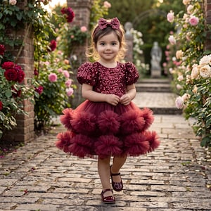 Op de afbeelding: Een jong meisje in een bordeauxrood paillettenjurk met een gelaagde tule rok en bijpassende haarband en schoenen. De jurk heeft pofmouwen. Ze loopt op een geplaveide weg omringd door bloemen.