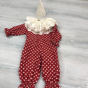 May include: A red and white polka dot clown costume with a lace collar and a matching cone hat. The long-sleeved jumpsuit has a ruffled hem and cuffs. The hat is a cream color.