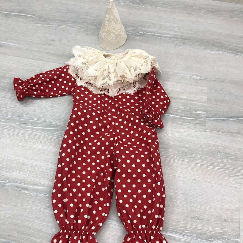 May include: A red and white polka dot clown costume with a lace collar and a matching cone hat. The long-sleeved jumpsuit has a ruffled hem and cuffs. The hat is a cream color.