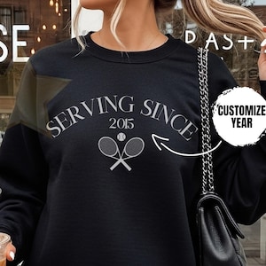 May include: Black sweatshirt with the text "SERVING SINCE 2015" and tennis rackets. The sweatshirt has a crew neck and long sleeves. The words "CUSTOMIZE YEAR" are also visible. The sweatshirt is part of a tennis-themed apparel collection.