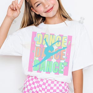 May include: A person wearing a white t-shirt with the word "DANCE" in a pink and green design. The shirt also features a teal dancer silhouette. The person is wearing pink heart-shaped sunglasses and pink and white checkered shorts.