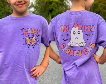 Puede incluir: Dos chicas con camisetas moradas con temática de Halloween. La camiseta delantera dice "Ruby" con un lazo rosa y una telaraña. La camiseta trasera dice "In My Spooky Era" con un fantasma con un lazo rosa.
