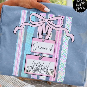 Op de afbeelding: Een blauw t-shirt met een afbeelding van een roze strik bevestigd aan twee op elkaar gestapelde rechthoekige badges. De bovenste badge toont de naam Savannah in cursief schrift en de onderste badge bevat de tekst Medical Assistant in zwart.
