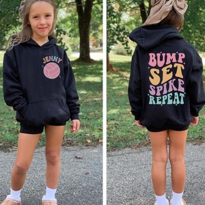 May include: Black hooded sweatshirt with a volleyball graphic and the name "Jenny" on the front. The back of the sweatshirt has the text "Bump Set Spike Repeat" in colourful retro lettering.