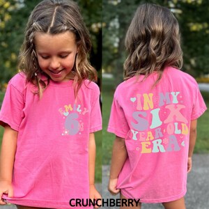 May include: Pink t-shirt with a colourful graphic that says "Emily 6" on the front and "In My Six Year Old Era" on the back.
