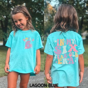 May include: A light blue t-shirt with a colourful graphic that says "In My Six Year Old Era". The shirt is shown on a child.