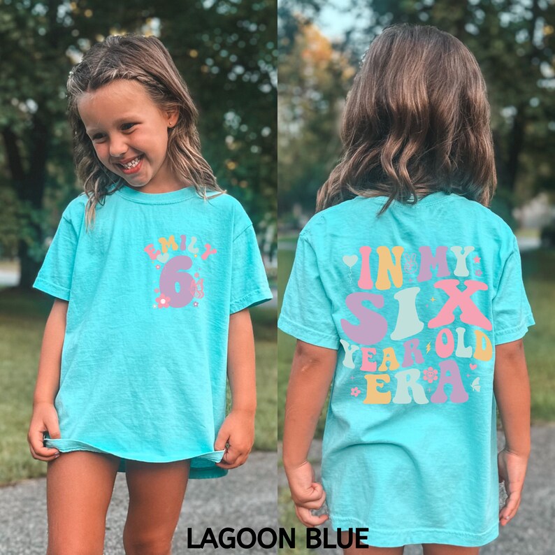 May include: A light blue t-shirt with a colourful graphic that says "In My Six Year Old Era". The shirt is shown on a child.