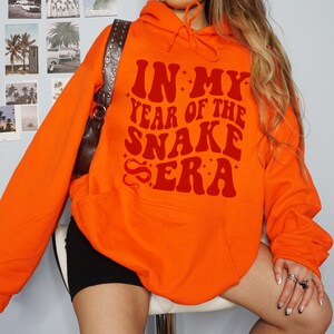 May include: Orange hoodie with the text "In My Year of the Snake Era" in red.