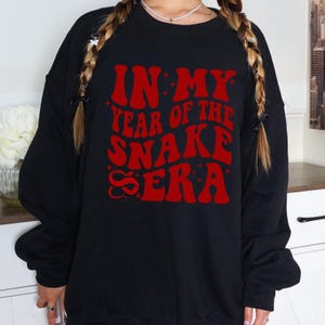 May include: Black sweatshirt with red text that reads "In My Year of the Snake Era" with a snake graphic.