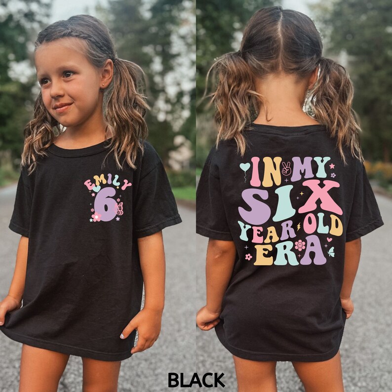 May include: A black t-shirt with a colourful graphic that says "In My Six Year Old Era" on the back and "Emily 6" on the front. The graphic features a rainbow colour scheme with flowers and a butterfly.