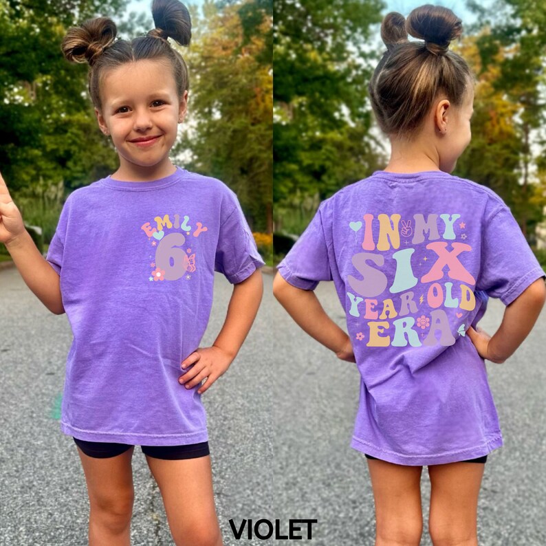 May include: Purple short-sleeved t-shirt with a colourful graphic that says "Emily 6" on the front and "In My Six Year Old Era" on the back.