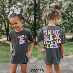 May include: A dark grey t-shirt with a colourful graphic that says "In My Six Year Old Era" on the back. The front of the shirt has a pink number 6 with flowers and the name "Emily" above it.