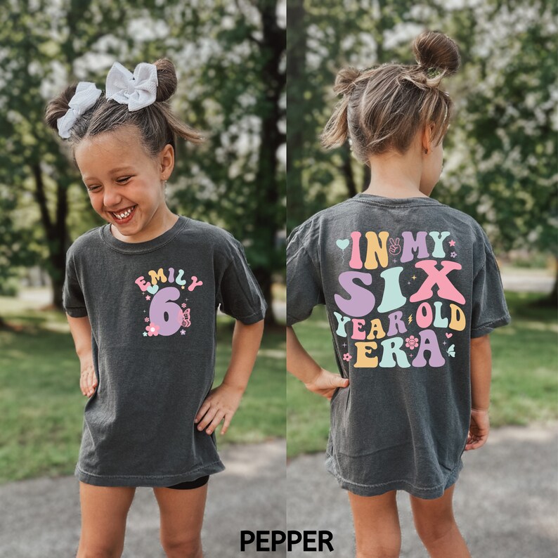May include: A dark grey t-shirt with a colourful graphic that says "In My Six Year Old Era" on the back. The front of the shirt has a pink number 6 with flowers and the name "Emily" above it.