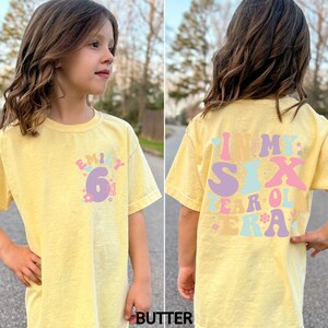 May include: A yellow t-shirt with a colourful graphic that says "Emily 6" on the front and "In My Six Year Old Era" on the back.