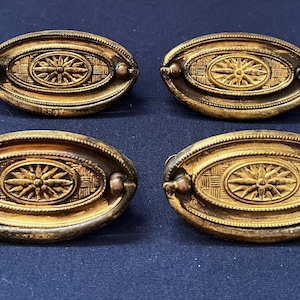 Może przedstawiać: Cztery antyczne mosiężne uchwyty do szafek w kształcie owalu z ozdobnym wzorem gwiazdy w środku. Każdy uchwyt ma dwa małe miedziane śruby z tyłu.