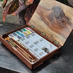 May include: A wooden watercolor paint set with a hinged lid. The set includes a tray of watercolor paints, a tray of empty wells, and a set of brushes. The set is open and the lid is propped up.