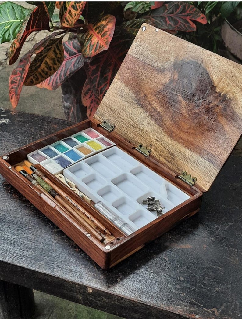 May include: A wooden watercolor paint set with a hinged lid. The set includes a tray of watercolor paints, a tray of empty wells, and a set of brushes. The set is open and the lid is propped up.