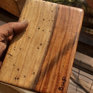 May include: A wooden cutting board with a natural wood grain finish. The board has a dark brown stain on one side and is marked with the word "SUMS".