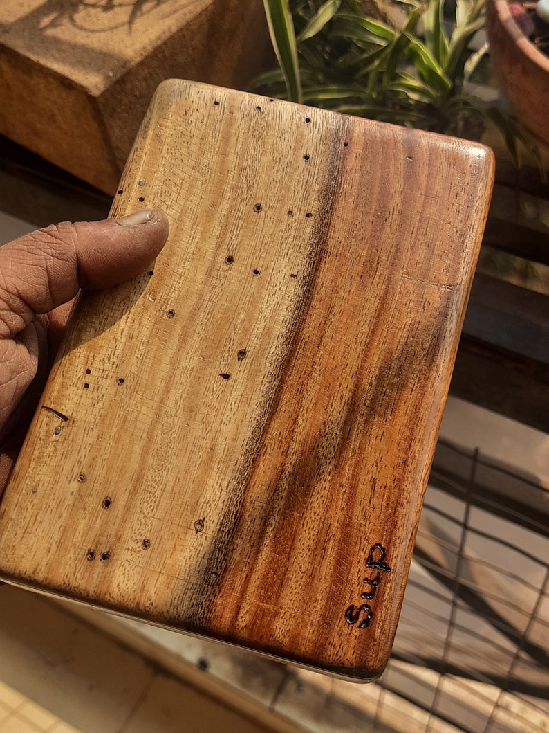 May include: A wooden cutting board with a natural wood grain finish. The board has a dark brown stain on one side and is marked with the word "SUMS".