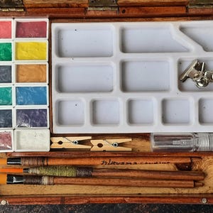 May include: A wooden watercolor paint box with a white plastic tray, 12 paint pans, 3 paintbrushes, 2 clothespins, a small spray bottle, and the text "Irisvana Creations".