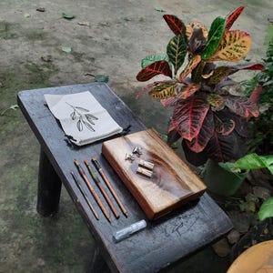May include: A wooden box with a natural wood finish and a clear epoxy resin top. The box is open and contains a set of paintbrushes and a small bag with a leaf design. The box is sitting on a dark wooden table.