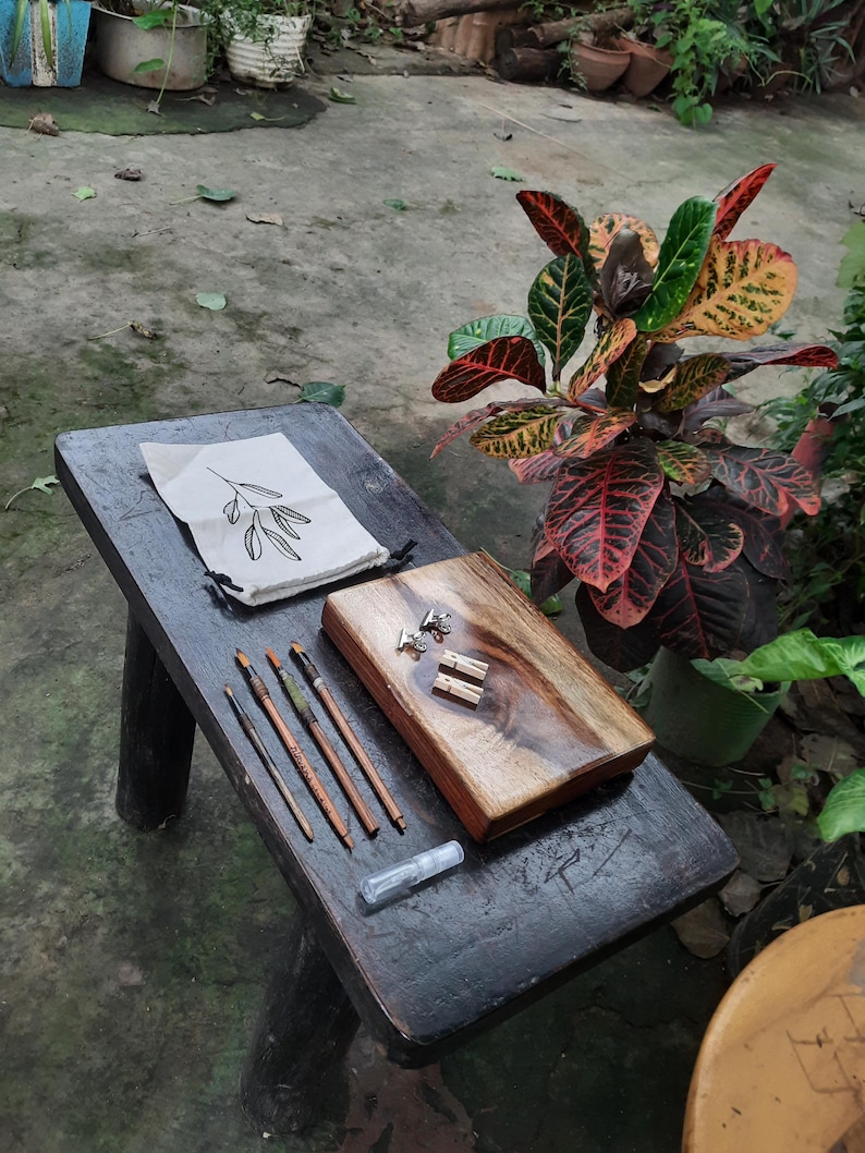 May include: A wooden box with a natural wood finish and a clear epoxy resin top. The box is open and contains a set of paintbrushes and a small bag with a leaf design. The box is sitting on a dark wooden table.