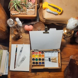 May include: A wooden watercolor travel box featuring a hinged lid with a metal clip, a grid of sixteen circular paint pans in various colors, and a white mixing tray containing a black binder clip and two small wooden clothespins.
