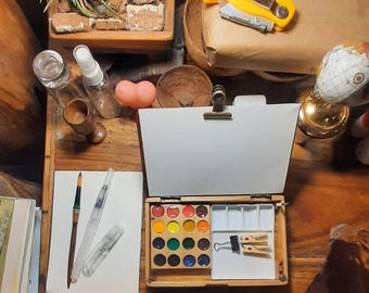May include: A wooden watercolor travel box featuring a hinged lid with a metal clip, a grid of sixteen circular paint pans in various colors, and a white mixing tray containing a black binder clip and two small wooden clothespins.