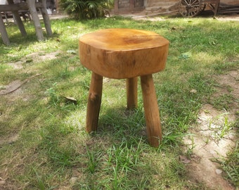 May include: A rustic wooden stool with a natural wood finish and three legs. The stool is made from a single piece of wood and has a unique, organic shape.
