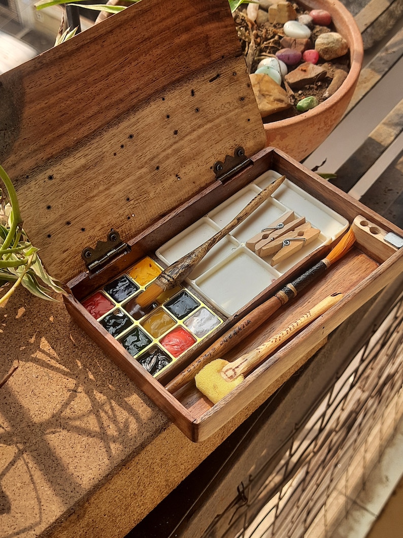 May include: A wooden watercolor paint set with a hinged lid. The set includes a palette of 12 watercolor paints, two paintbrushes, and two clothespins. The paint set is open and the lid is propped up.