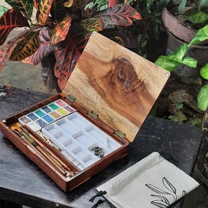 May include: A wooden watercolor paint box with a lid open, revealing a set of watercolor paints, brushes, and a white plastic tray with empty wells. The box is sitting on a dark wooden surface, and a small white canvas bag with a black drawstring is lying next to it.