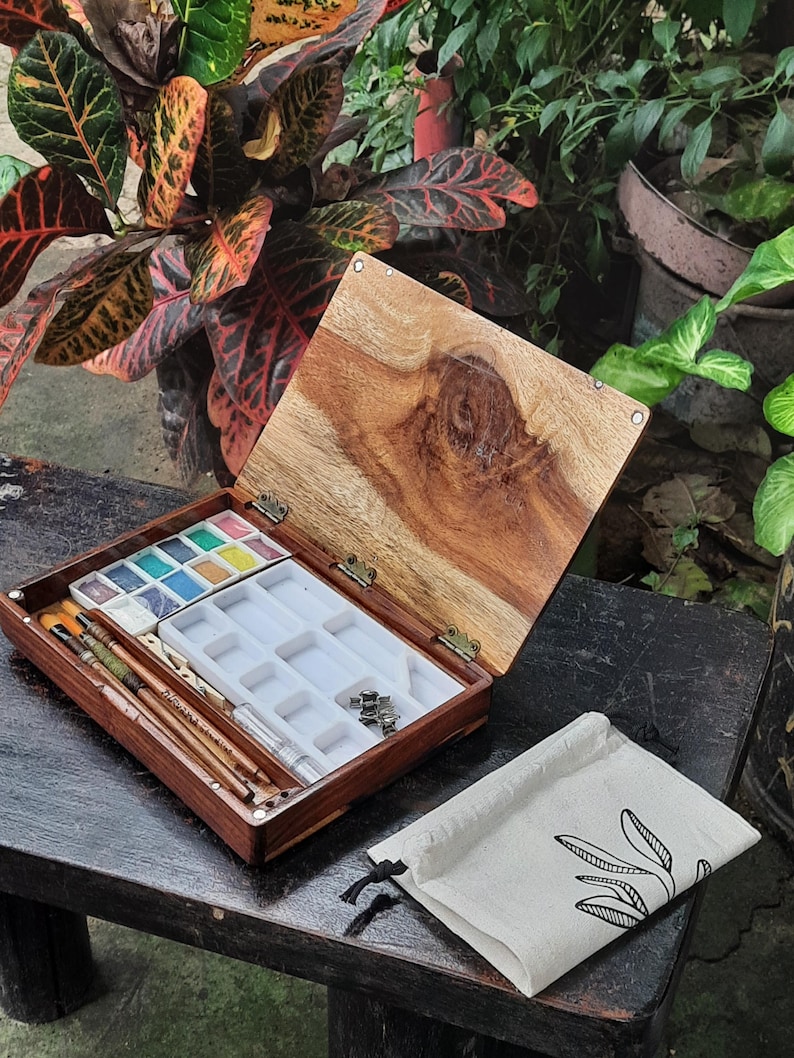 May include: A wooden watercolor paint box with a lid open, revealing a set of watercolor paints, brushes, and a white plastic tray with empty wells. The box is sitting on a dark wooden surface, and a small white canvas bag with a black drawstring is lying next to it.
