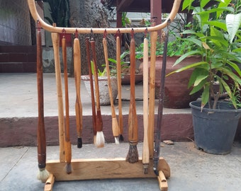 Puede incluir: Un estante de madera para pinceles que muestra varios pinceles de caligrafía. El estante está hecho de madera clara con una parte superior curvada y una base rectangular. Los pinceles tienen mangos de madera y cerdas de diferentes colores, colgando del estante.