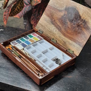 May include: A wooden watercolor paint box with a lid open, revealing a tray of watercolor pans, brushes, and a small bottle of water. The box is made of dark wood with a light wood interior.
