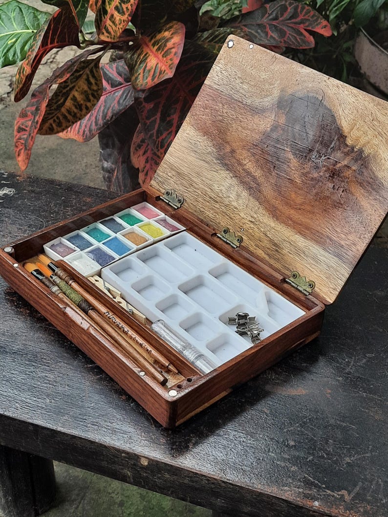 May include: A wooden watercolor paint box with a lid open, revealing a tray of watercolor pans, brushes, and a small bottle of water. The box is made of dark wood with a light wood interior.