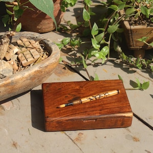 Könnte beinhalten: Eine dunkelbraune Holzbox mit einem Stift darauf, auf einer verwitterten Oberfläche. Der Stift hat einen goldfarbenen Schaft mit einem dekorativen Muster. Die Szene wird durch Grün und eine rustikale Holzschale ergänzt.
