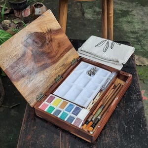 May include: A wooden watercolor paint set with a hinged lid, containing a palette of 12 watercolor paints, a white plastic tray with 12 wells, a small white plastic container, a white plastic clothespin, and a set of brushes. The set is open and sitting on a wooden table.
