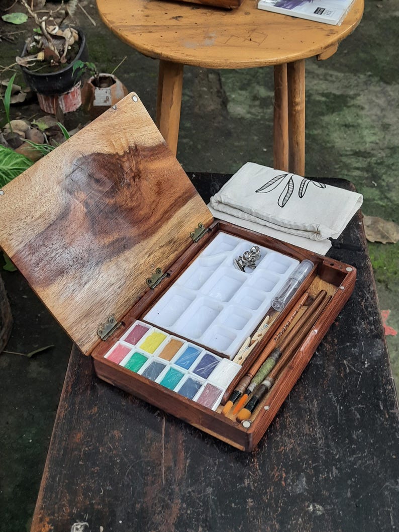 May include: A wooden watercolor paint set with a hinged lid, containing a palette of 12 watercolor paints, a white plastic tray with 12 wells, a small white plastic container, a white plastic clothespin, and a set of brushes. The set is open and sitting on a wooden table.