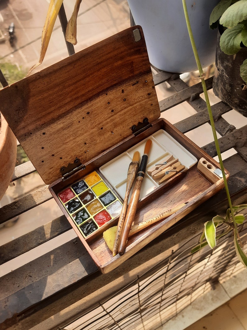 May include: A wooden watercolor paint set with a hinged lid. The set includes a palette of 12 watercolor paints, two paintbrushes, a sponge, and two clothespins.