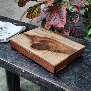 May include: A rectangular wooden cutting board with a natural wood finish and a dark brown border. The board has a unique grain pattern and is sitting on a dark wooden surface.