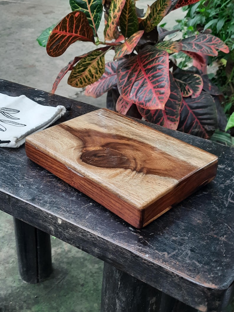 May include: A rectangular wooden cutting board with a natural wood finish and a dark brown border. The board has a unique grain pattern and is sitting on a dark wooden surface.