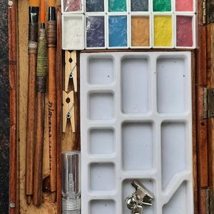 May include: A wooden watercolor paint set with a variety of colors, including red, green, blue, yellow, and brown. The set includes three brushes, a small bottle of water, and a palette with multiple compartments.