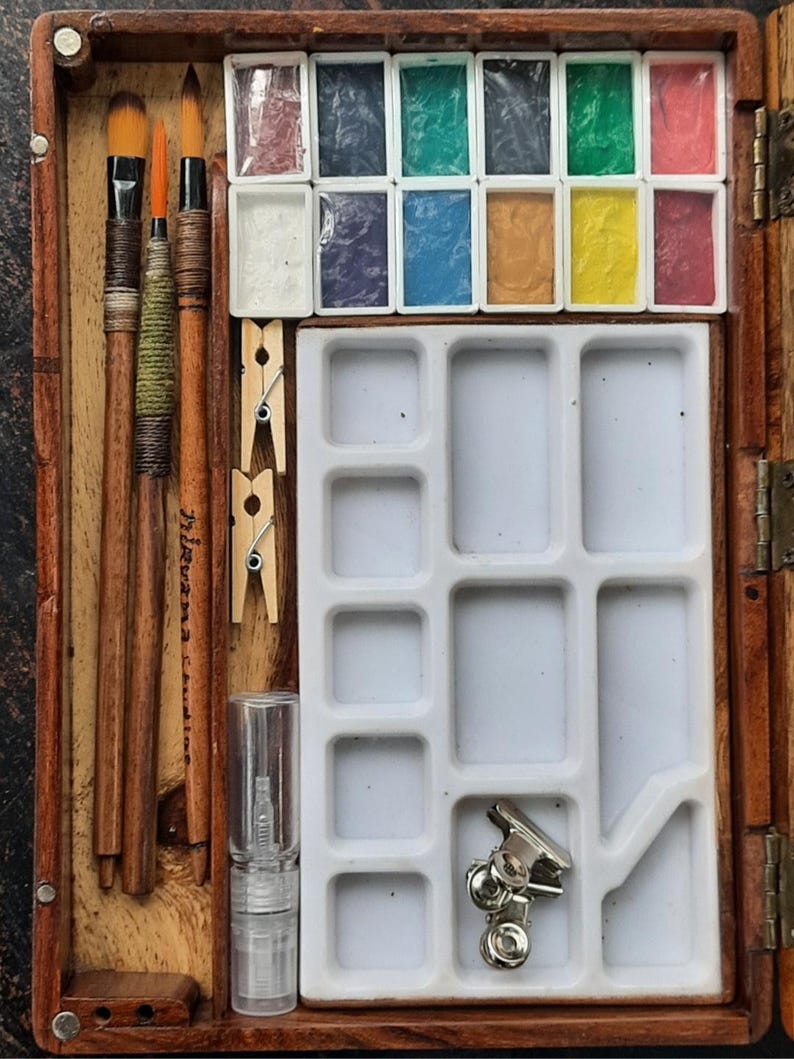 May include: A wooden watercolor paint set with a variety of colors, including red, green, blue, yellow, and brown. The set includes three brushes, a small bottle of water, and a palette with multiple compartments.