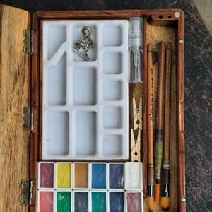 May include: A wooden box with a hinged lid, containing a watercolor paint set. The set includes 12 watercolor pans, two brushes, a small bottle of water, and a clip. The box is made of dark wood with a natural finish.