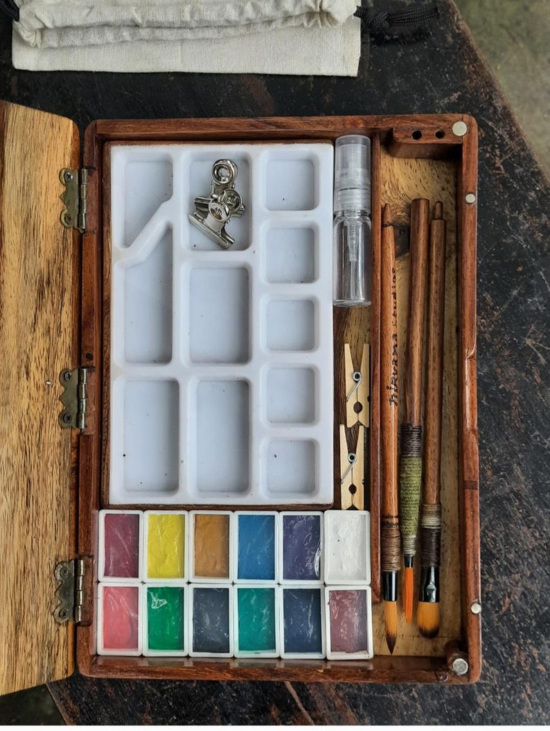 May include: A wooden box with a hinged lid, containing a watercolor paint set. The set includes 12 watercolor pans, two brushes, a small bottle of water, and a clip. The box is made of dark wood with a natural finish.