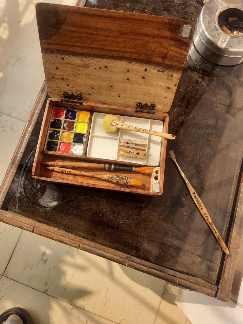 May include: A wooden box with a hinged lid, containing a watercolor paint set with a variety of colors, a sponge, and brushes. The box is open and the contents are visible.