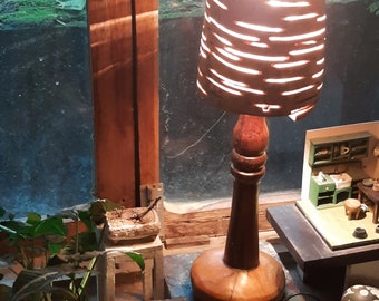 May include: A wooden table lamp with a unique, slatted, light brown shade. The lamp is lit and casting a warm glow. The lamp is on a wooden table with a blue and green painted surface.