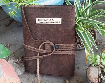 May include: A brown leather journal with a leather strap and buckle closure. The journal has a small, rectangular tag attached to the front with the text "Retroville & Nirvana Studios" written on it.