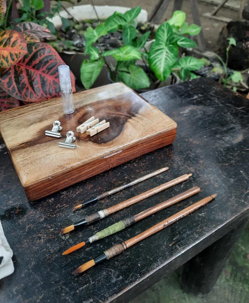 May include: A wooden box with a lid, two silver clips, three wooden clothespins, a small clear bottle, and four paintbrushes with wooden handles and colorful wrappings.
