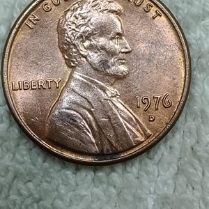 Könnte beinhalten: Eine kupferfarbene Ein-Cent-Münze aus den USA mit dem Profil von Abraham Lincoln, das nach rechts blickt. Der Text IN GOD WE TRUST ist oben gebogen, LIBERTY steht links und das Jahr 1976 mit dem Münzzeichen D befindet sich auf der rechten Seite.